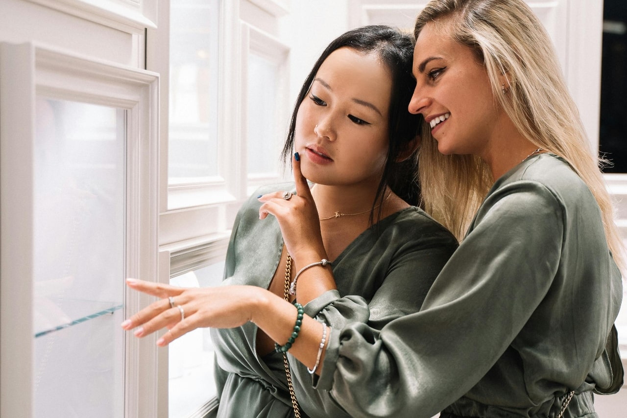 Two women wearing matching green silk robes, pointing at a jewelry display case.
