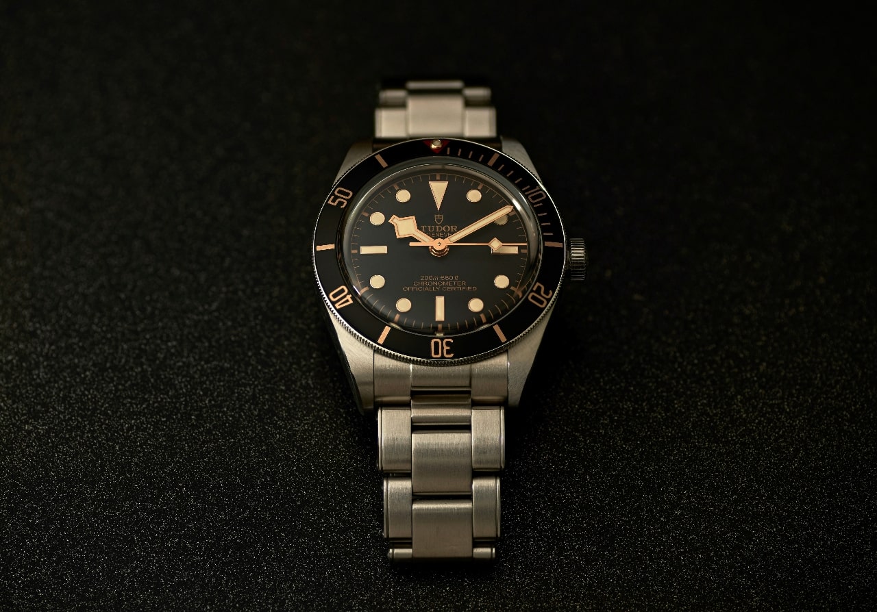 A TUDOR watch with a stainless steel bracelet and black dial on a black textured surface.