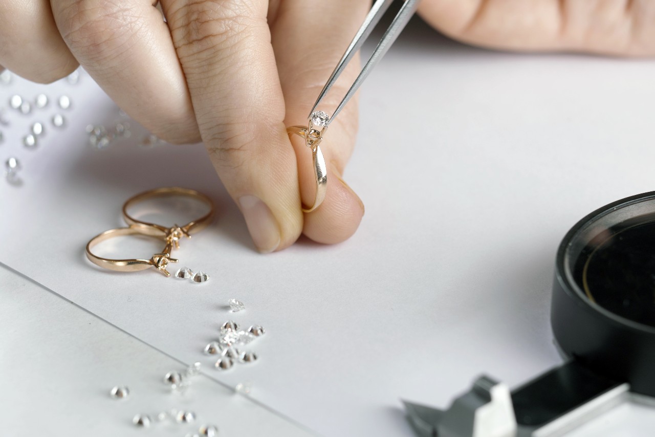 A jeweler sets a diamond into a gold ring using tweezers, surrounded by loose diamonds and ring settings.