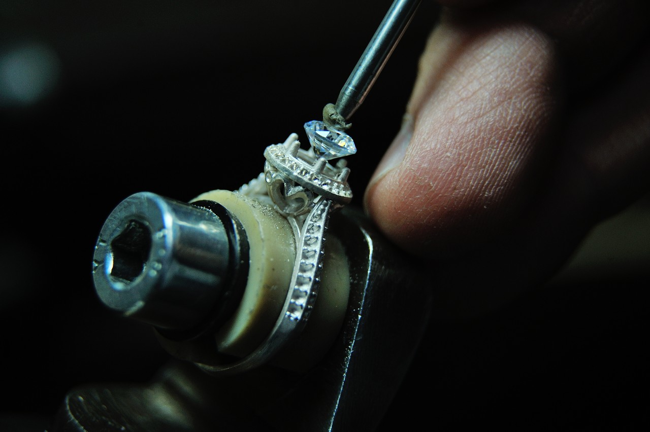 A close-up of a craftsman using a tool to secure a diamond in an intricate white gold engagement ring.