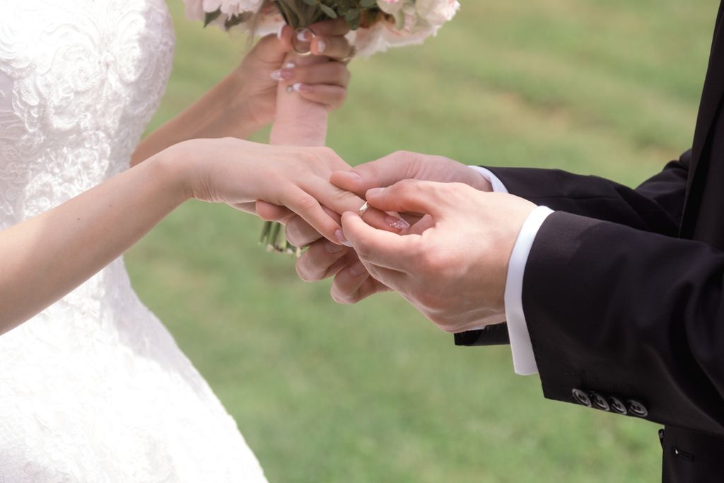 A groom places a ring on the bride’s finger outdoors, with the bride in a lace dress holding a bouquet and the groom in a black suit.