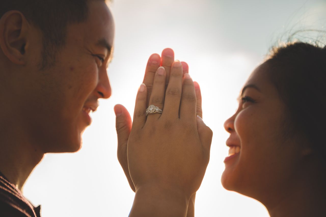 A smiling couple faces each other with their hands pressed together, showcasing a diamond engagement ring.