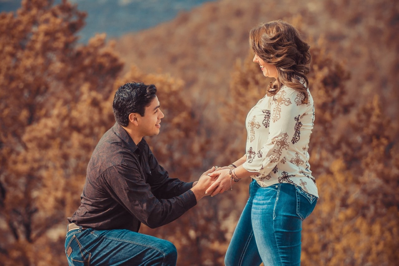 A man kneels outdoors amid autumn foliage, holding a woman’s hands in a romantic proposal.