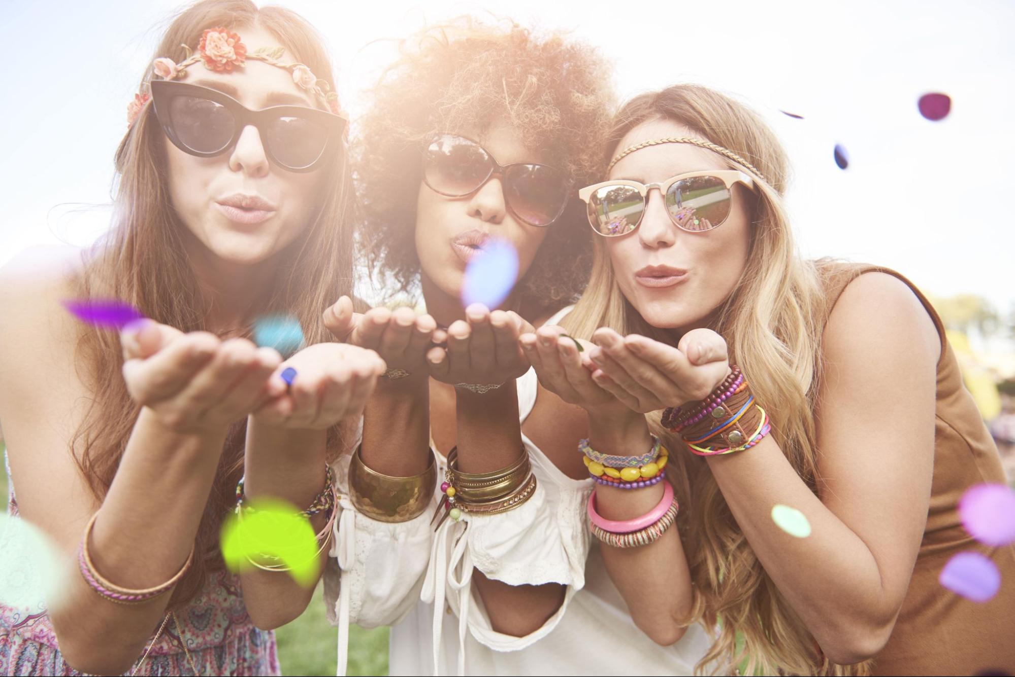 Three women with stacked bracelets all over their hands and sunglasses blow confetti towards the camera.