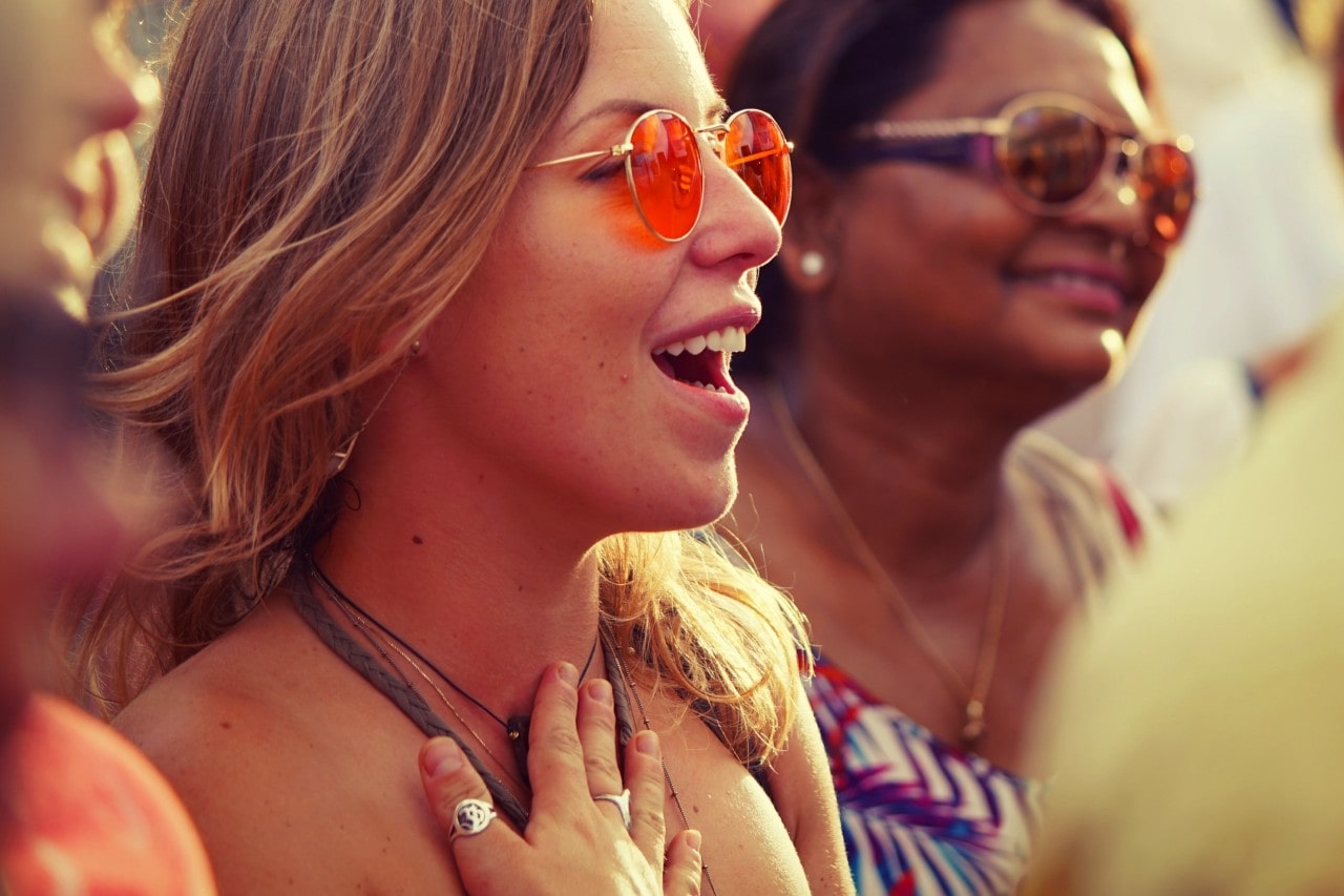A woman with sunglasses and stacked rings smiles while singing among other people.