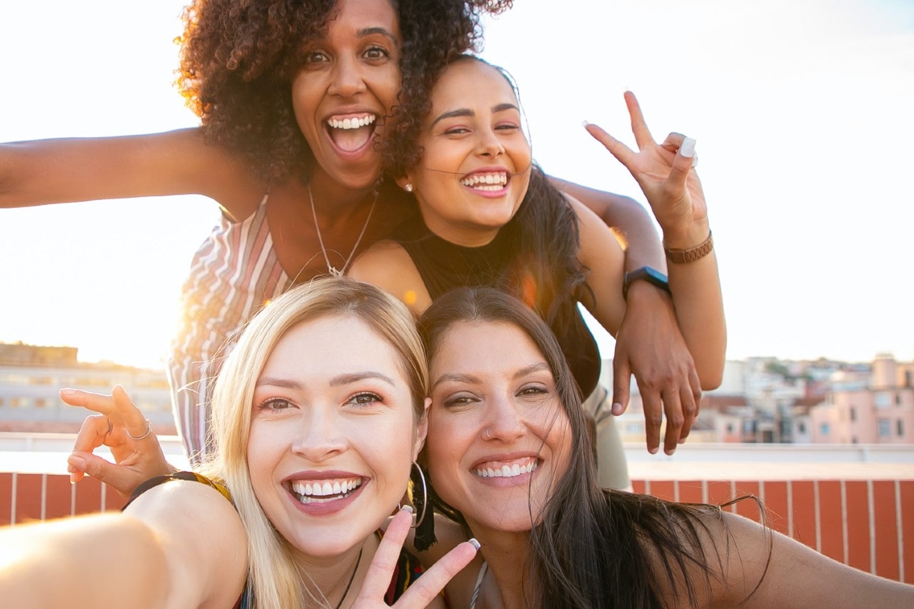 Four women smiling on a sunlight rooftop, wearing hoop earrings and gold necklaces.