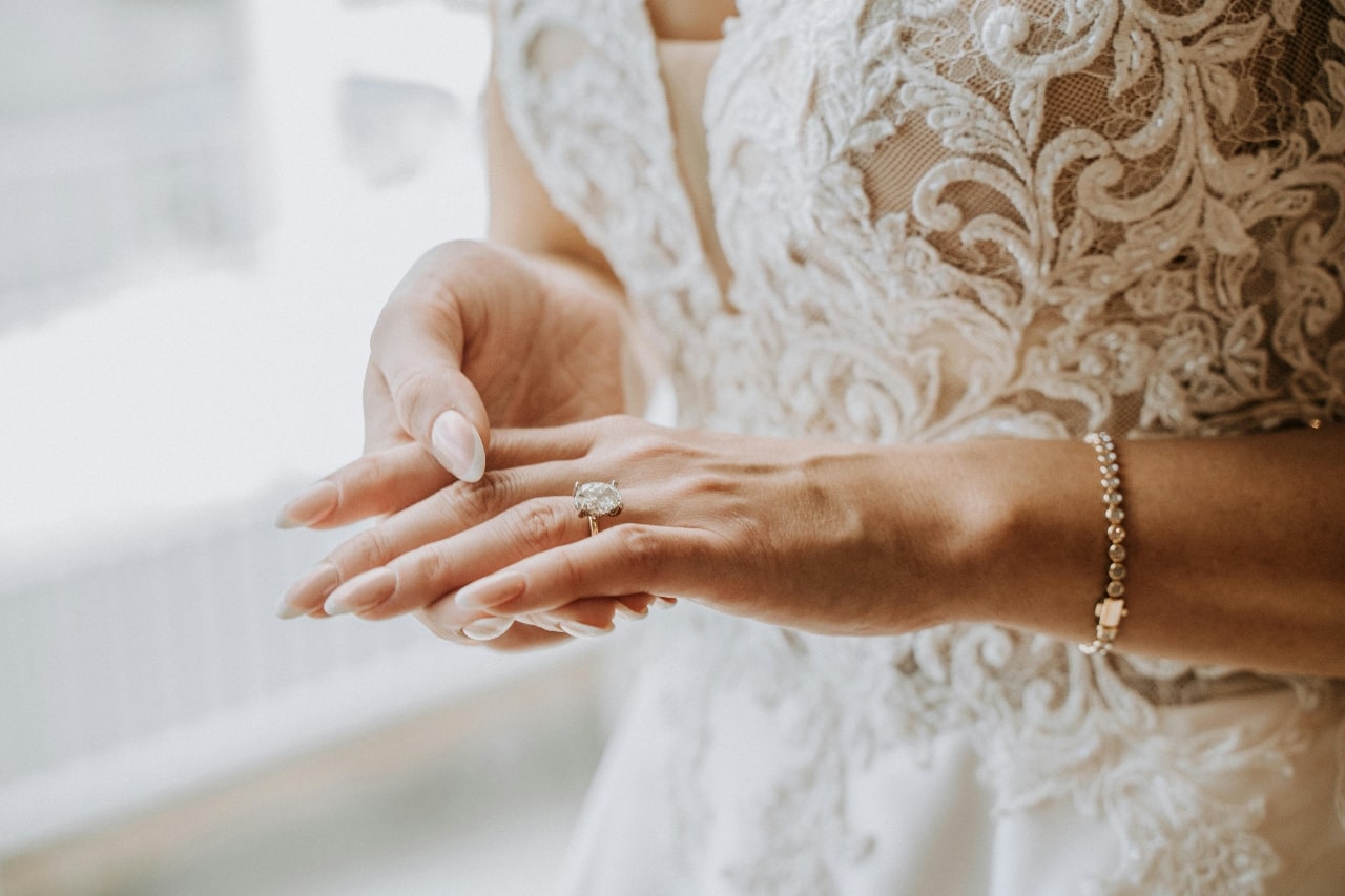 Bride in an elegant lace wedding gown gently adjusts a diamond engagement ring on her finger.