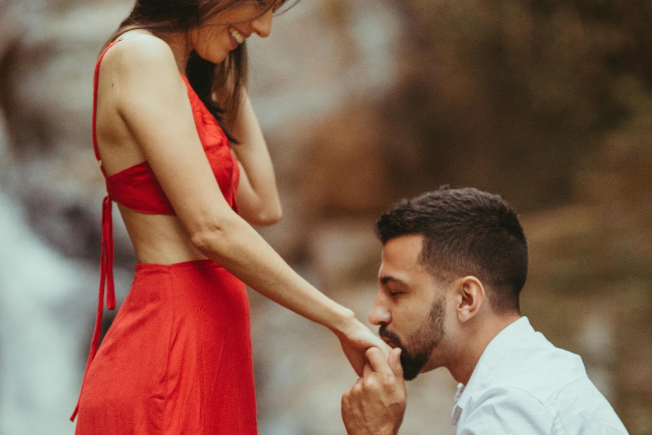 A man kneeling down proposing to a woman in a vibrant red dress against a blurred nature background.