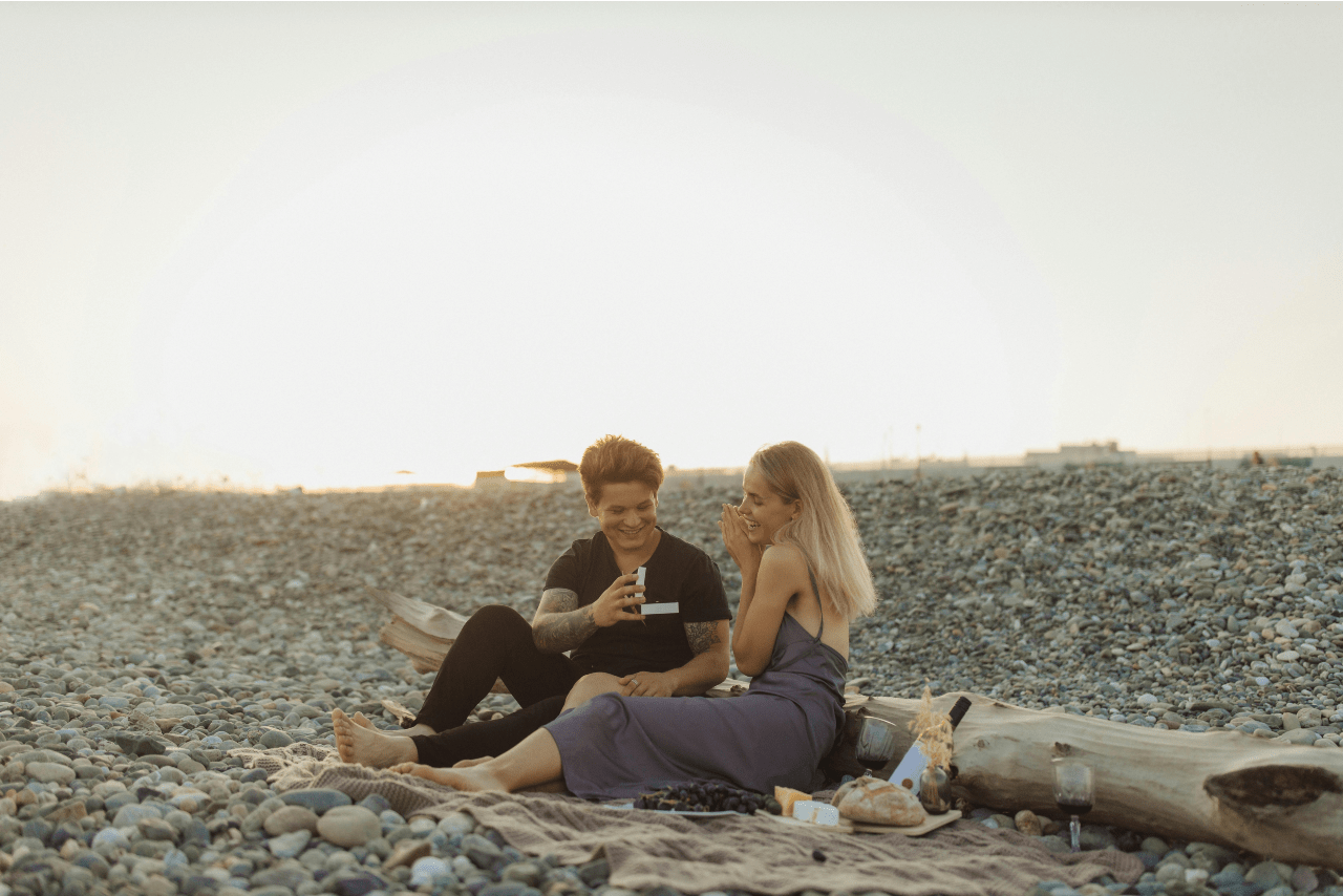 A man proposing with a ring box to a woman in a purple slip dress during a beach picnic.