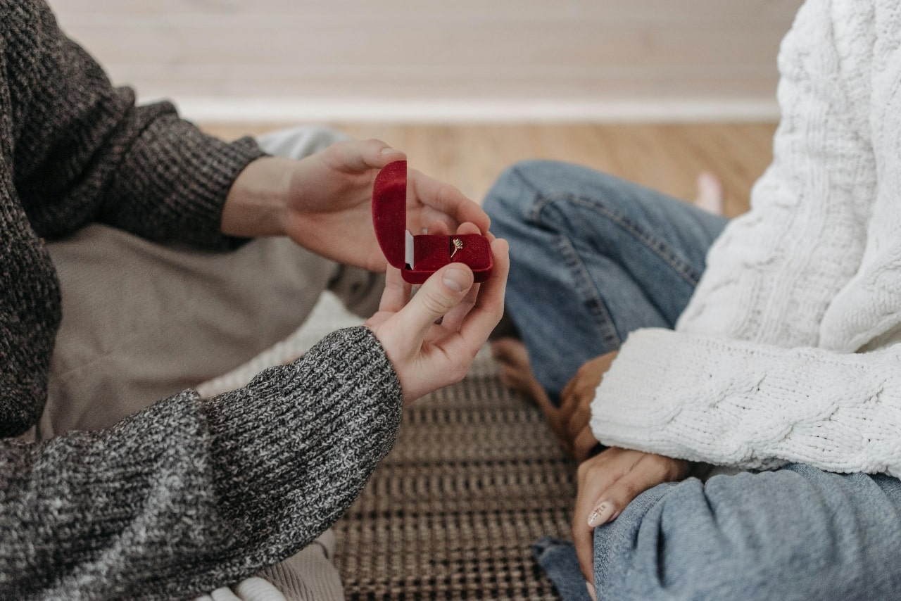 A cozy indoor marriage proposal with a couple sitting cross-legged on a rug while wearing knit sweaters.