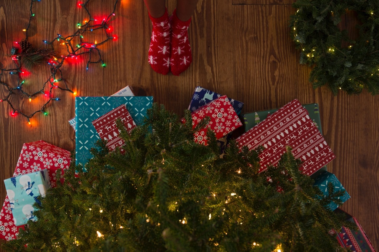 A top-down view of a person’s feet wearing cozy socks, standing before festively wrapped Christmas presents under a decorated tree, between colorful string lights and a holiday wreath.