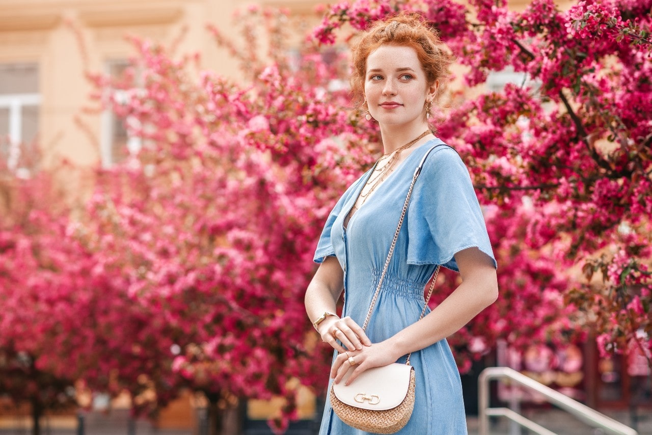 A ginger woman in a blue dress stands in front of blooming pink trees.