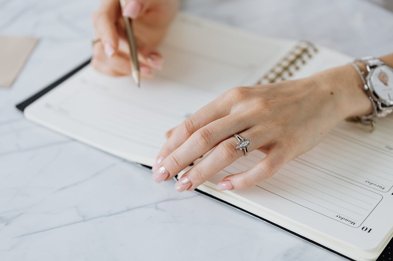 A close-up of hands writing in a notebook, showcasing a white gold marquise cut diamond engagement ring.