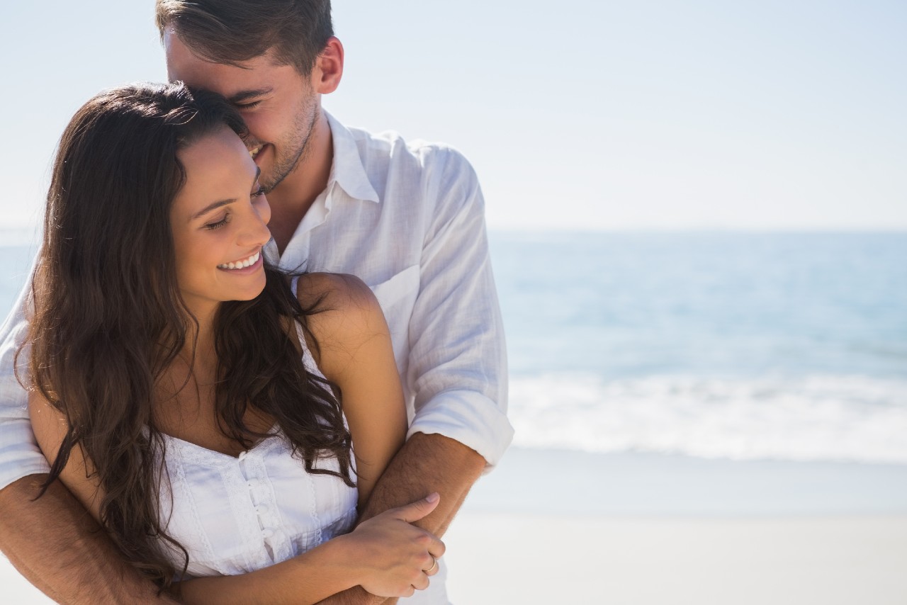 A man embracing his partner from behind while they both smile, set in front of the shoreline.