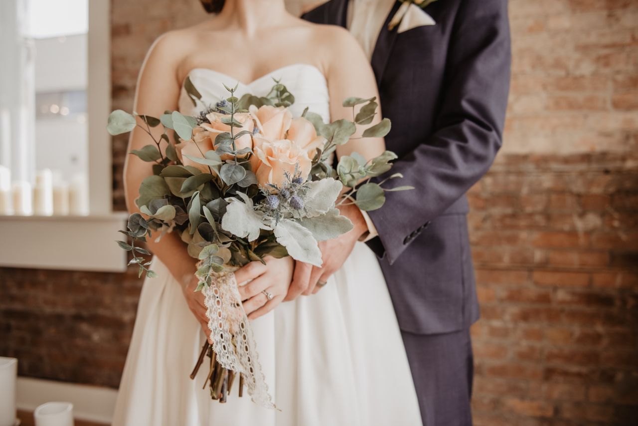 A bride standing in front of her groom and holding a beautiful wedding bouquet with light pink roses and some greenery, in front of a brick wall background.