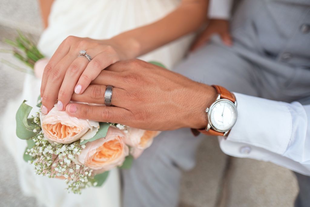 A freshly married couple still wearing their wedding attire and resting their hands together while showcasing a bouquet of light pink peonies and their shiny wedding ring and band.