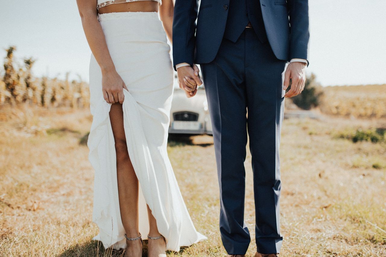 A newly married couple holding hands in their wedding party attire and standing in an open field in front of a vintage white car.