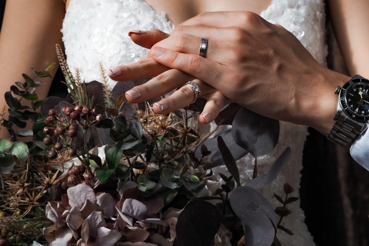 A bride in a wedding dress holding a fall-themed bouquet, with her husband's hand atop of hers and showcasing their wedding ring and band.