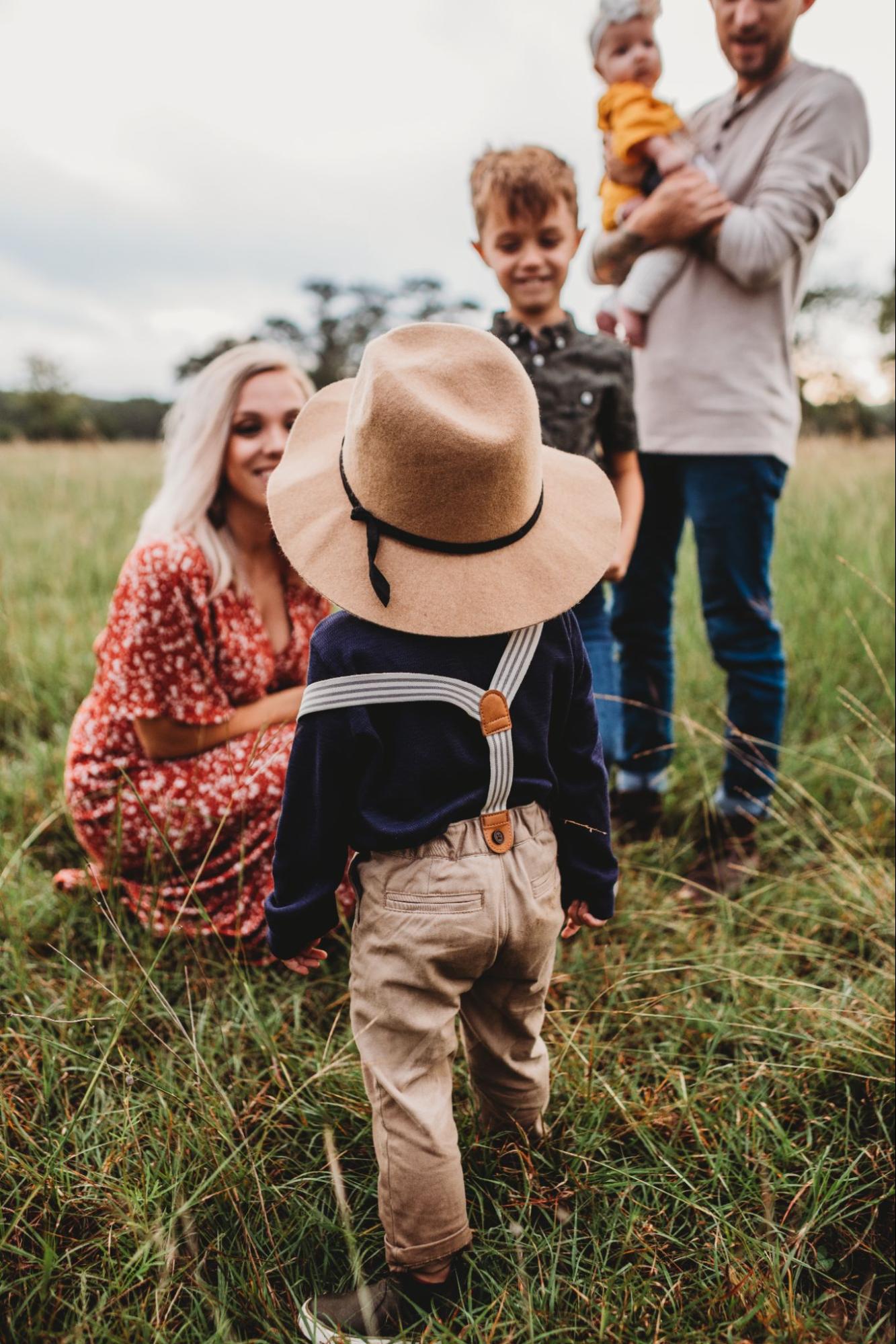 A toddler wearing a hat and suspenders walks through a grassy field toward smiling parents and siblings during a warm, outdoor family moment.