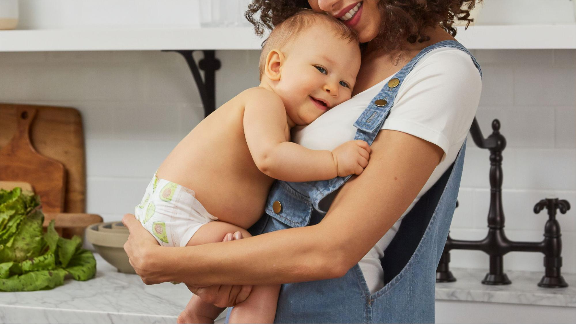 A close up of a mom with curly hair holding her baby in diapers in front of the kitchen counter.