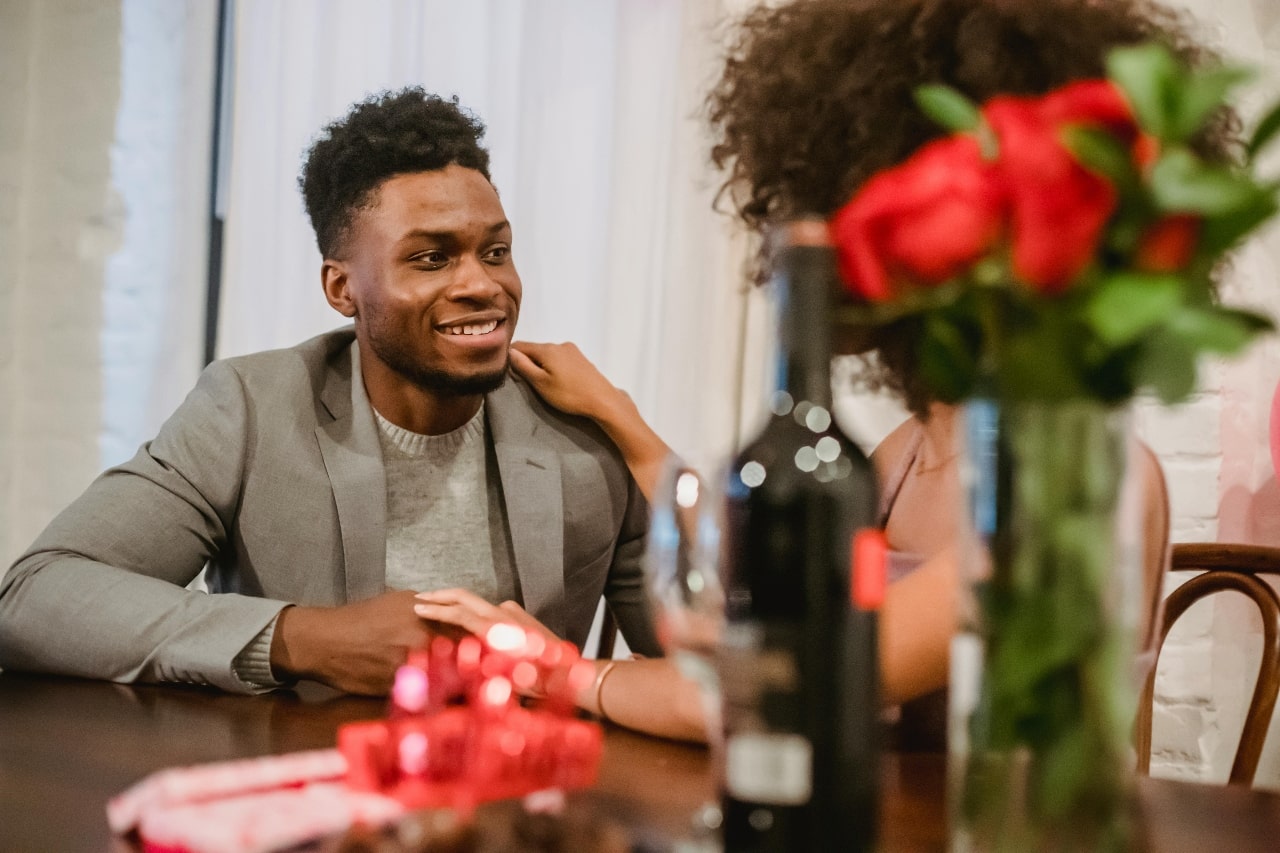 A couple sitting at the table with a red gift box on the table near a bottle of wine and red roses.