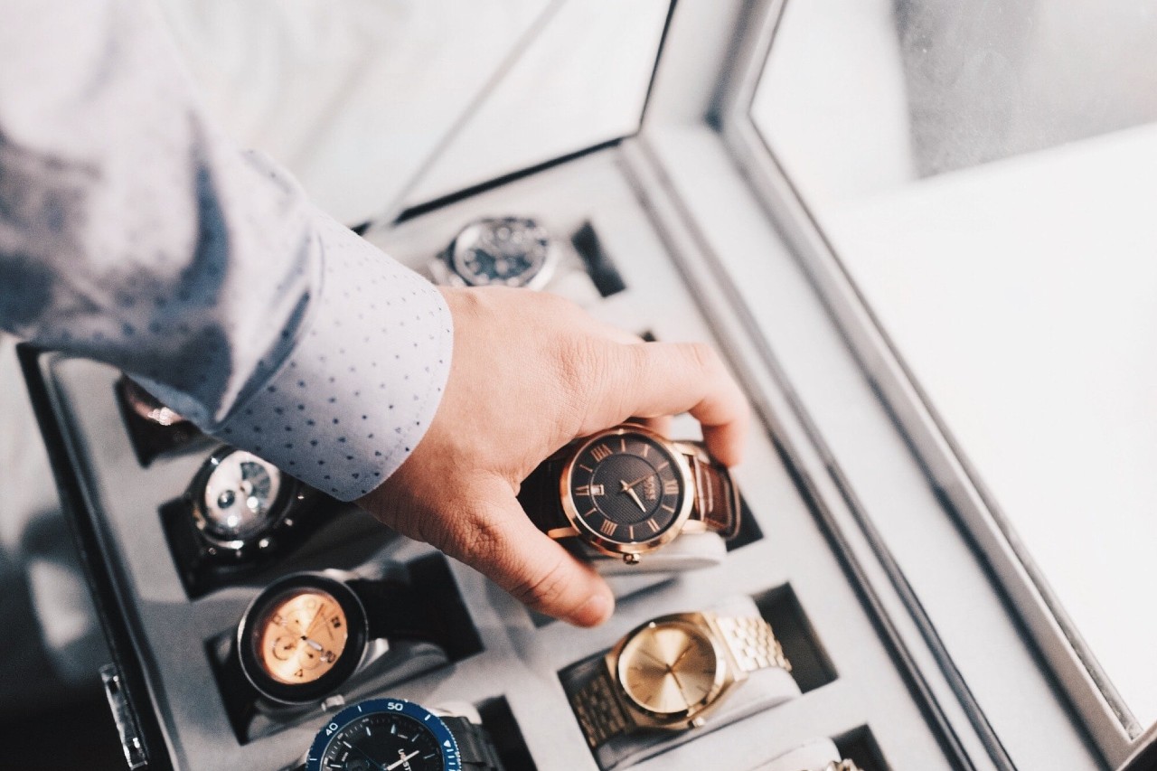 A person wearing a light patterned shirt selects a luxury wristwatch from a display case filled with various timepieces