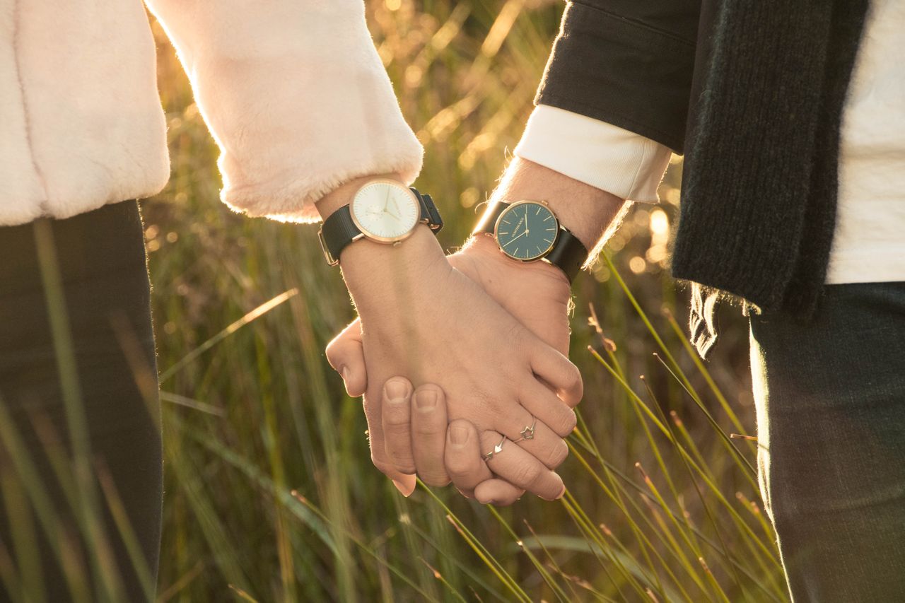 A close-up of a couple holding hands against a sunlight grass background, showcasing black leather wristwatches with white and black dials.