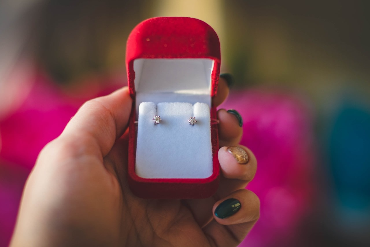 A manicured hand holding a red velvet earring box with diamond studs inside.