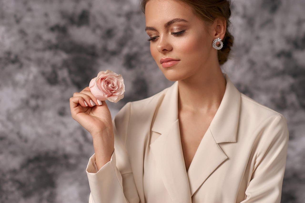 A woman in a cream blazer and sparkling earrings holding a pink rose against a soft gray background.