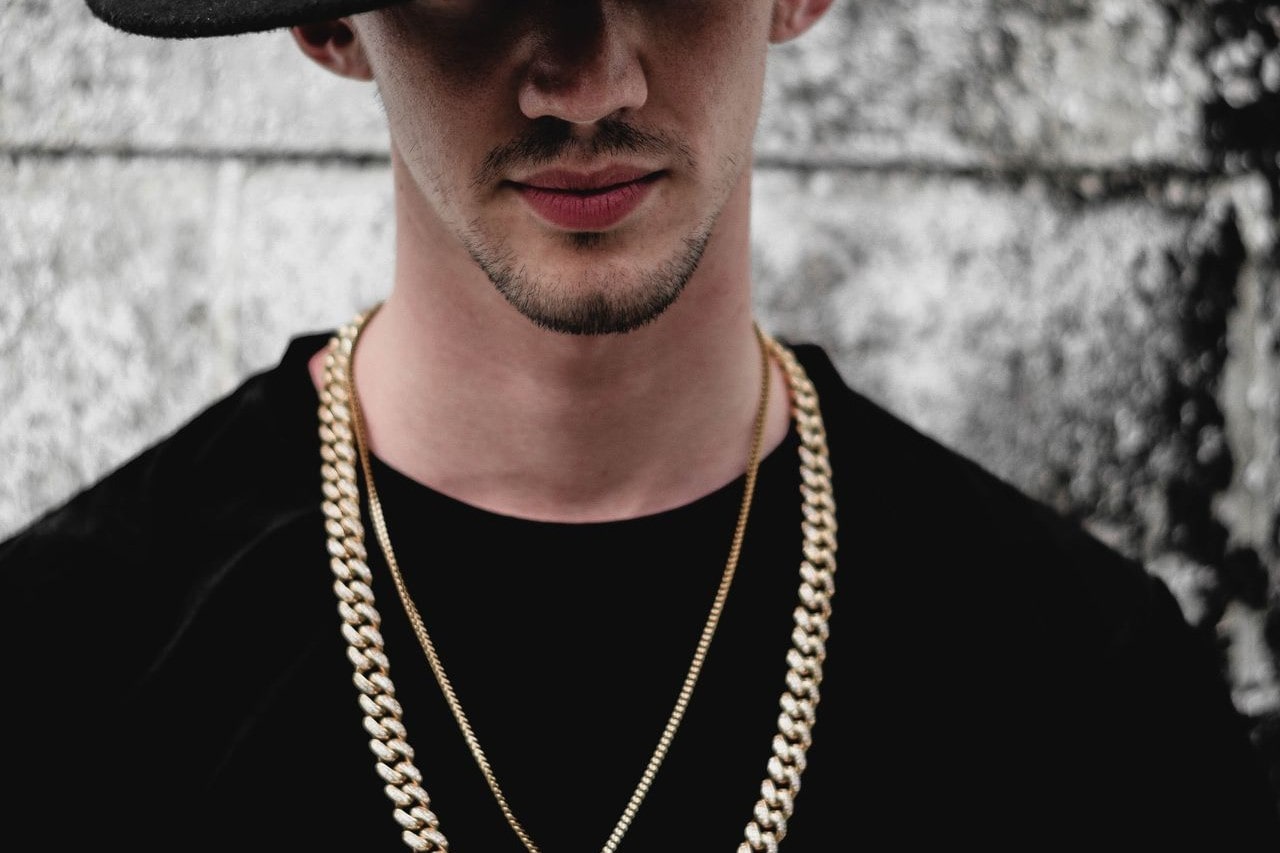 A man wearing a black shirt and a large black hat, wearing two golden chains of different thickness, in front of a grey background.