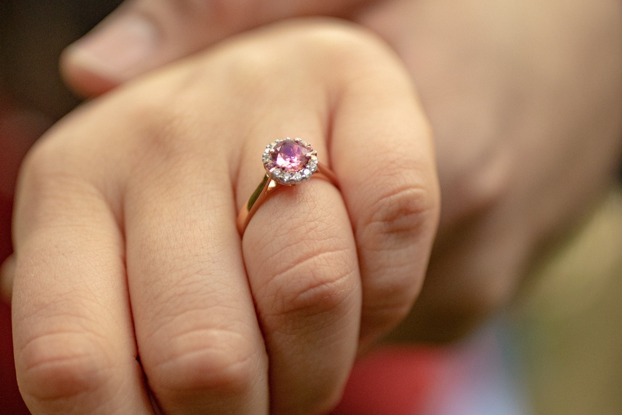 close up image of a hand wearing a rose gold engagement ring with a pink, halo set center stone