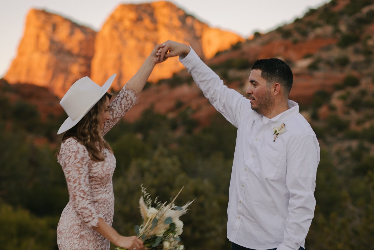 A couple, joyfully dancing outdoors against a backdrop of orange-tinted rocky cliffs. The woman wears a hat and holds flowers, the man in a white shirt.