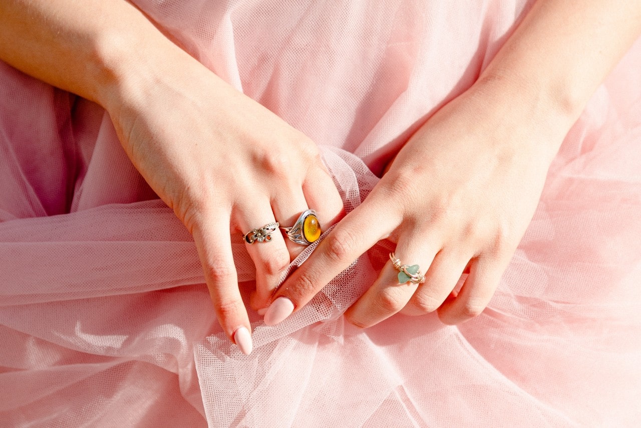 A close up of a woman’s hands bunching up her pink toile dress and showcasing her colorful rings.