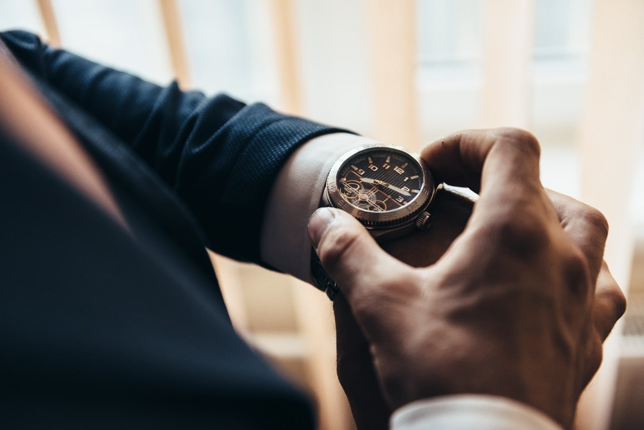 A man in a suit adjusts his luxury watch, displaying intricate gears and arab numerals.