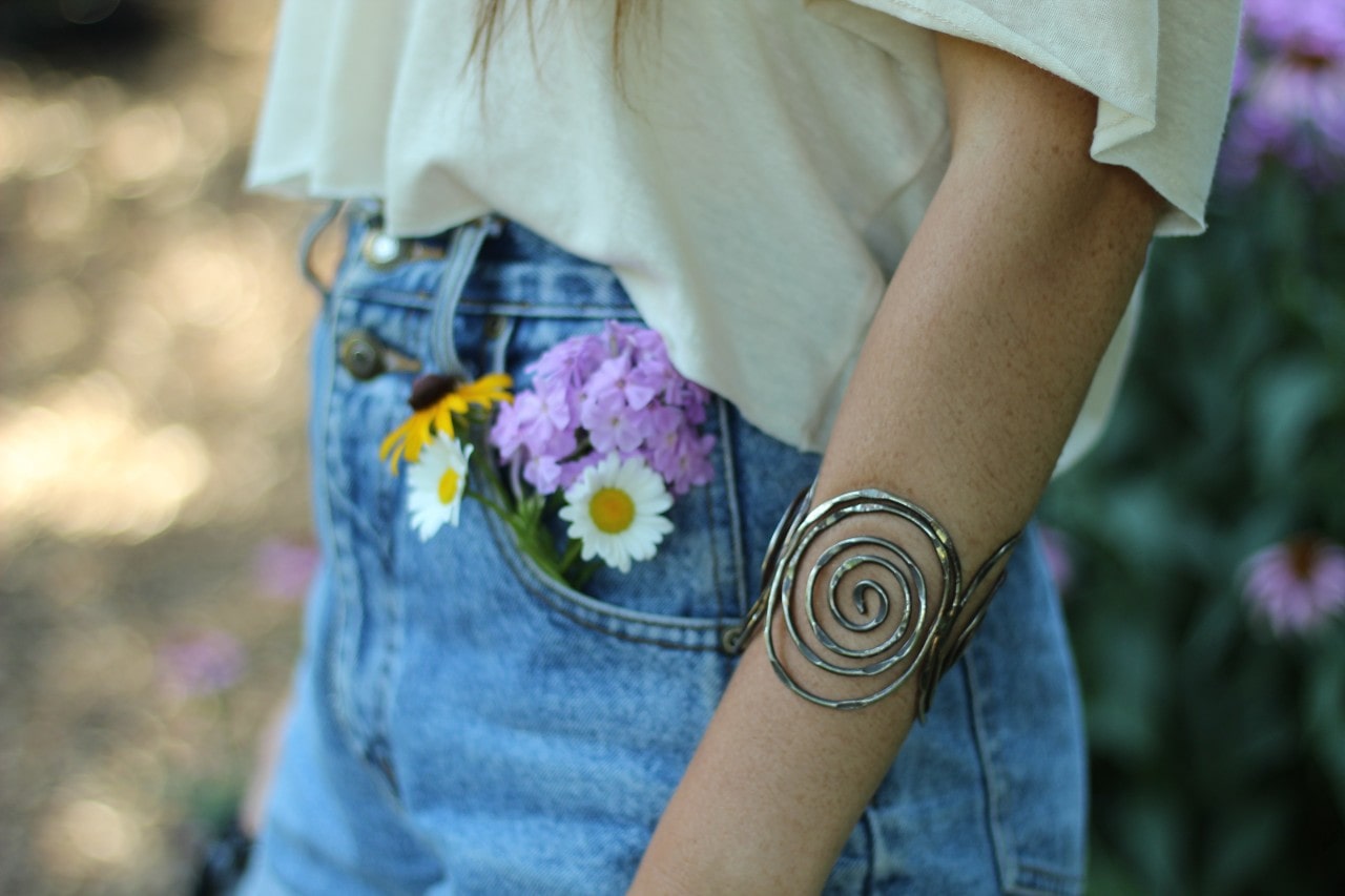 A close up of a person wearing a bold spiral metal cuff bracelet worn with a casual white top and denim jeans.