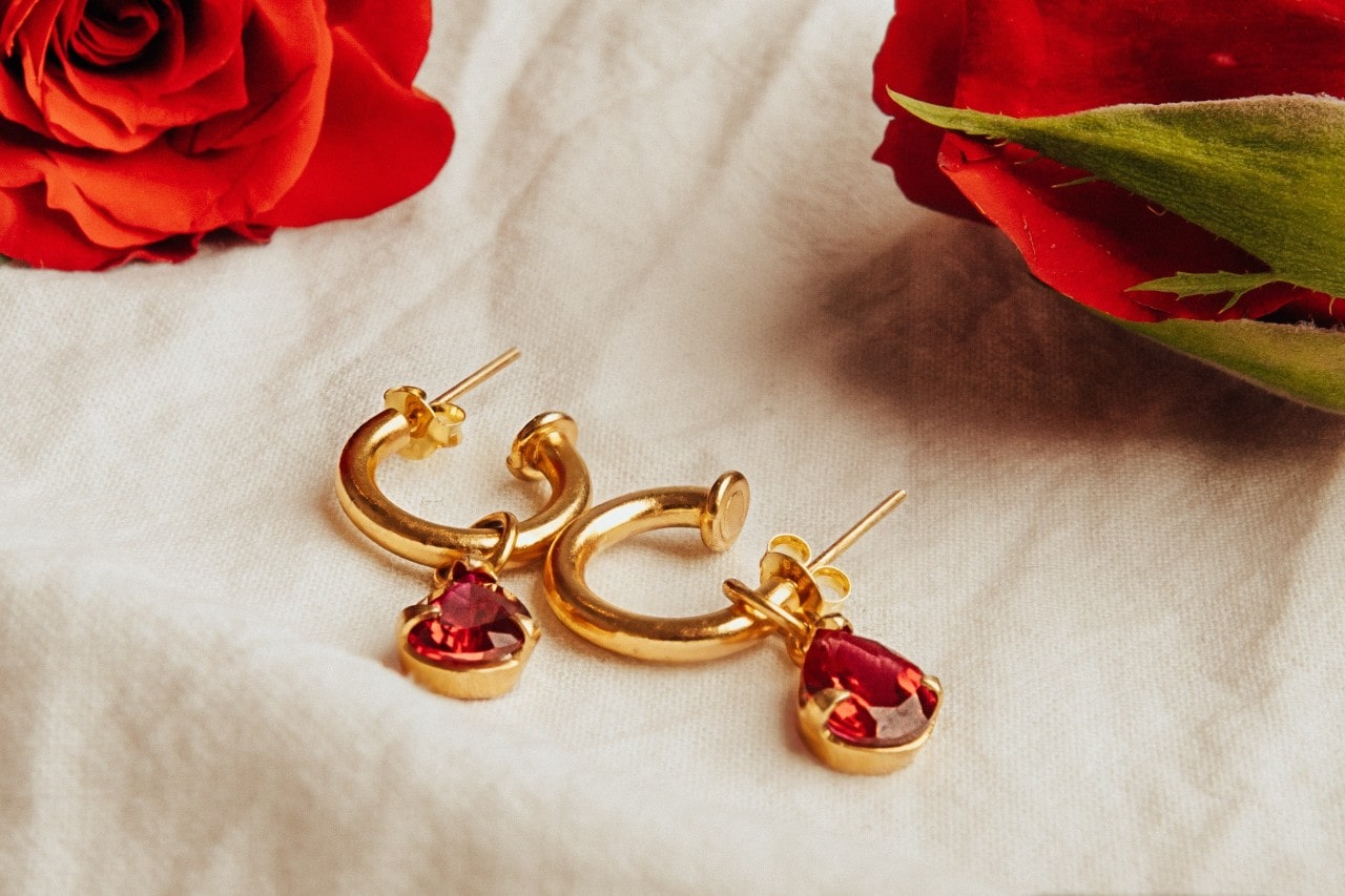 A yellow gold drop earrings with pear shaped rubies on a white cloth with two red roses nearby.