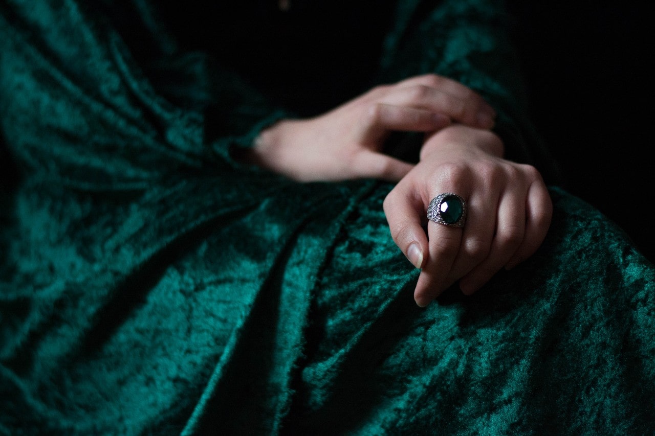A hand rests on a lap draped in rich green velvet, wearing a large emerald ring.