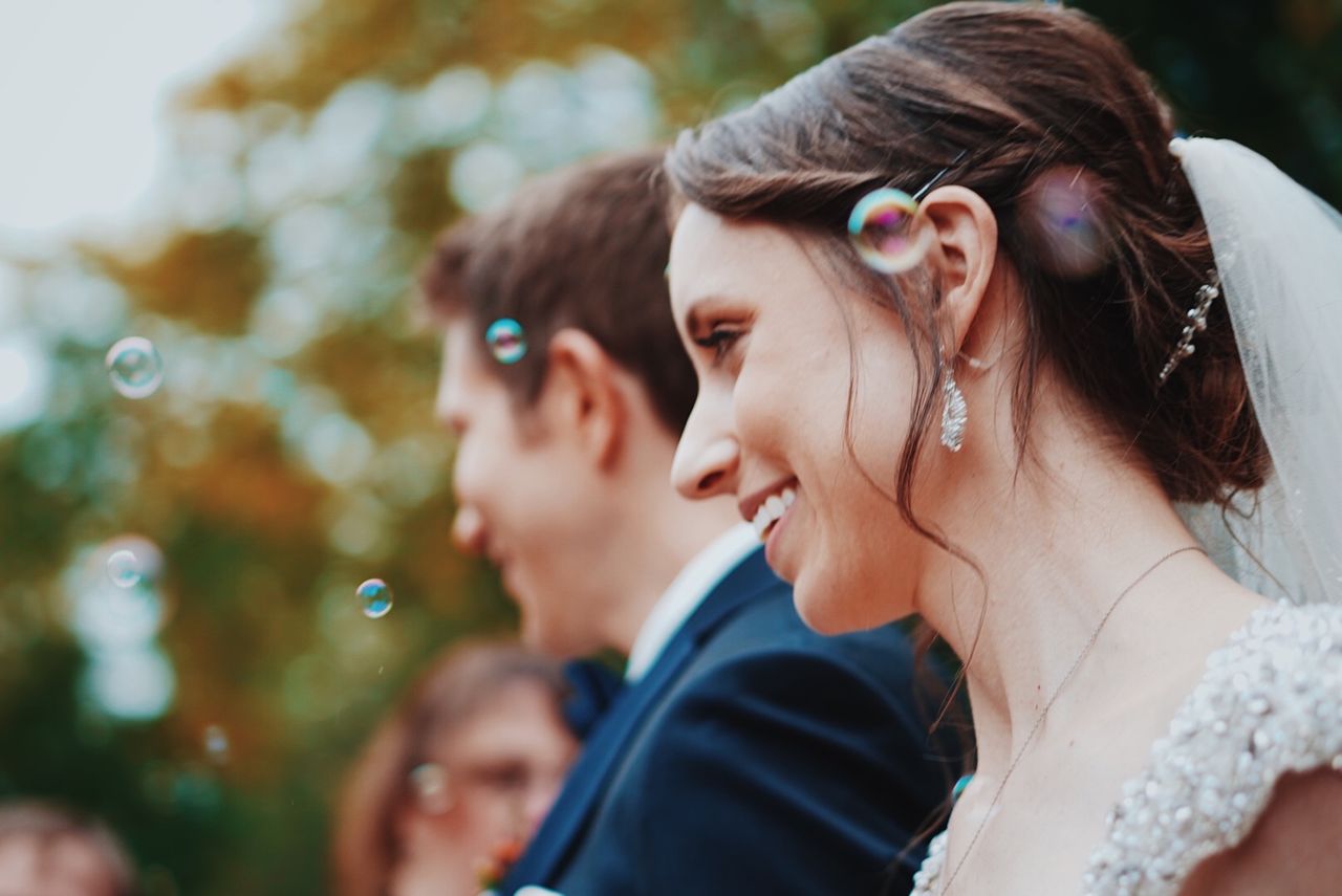 A smiling bride in a white gown and veil with sparkling earrings stands beside a groom in a suit.