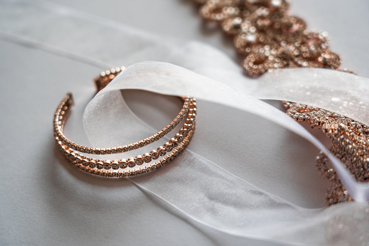 Elegant rose gold bracelet and tiara adorned with crystals, intertwined with a delicate sheer ribbon, lying on a soft white background.
