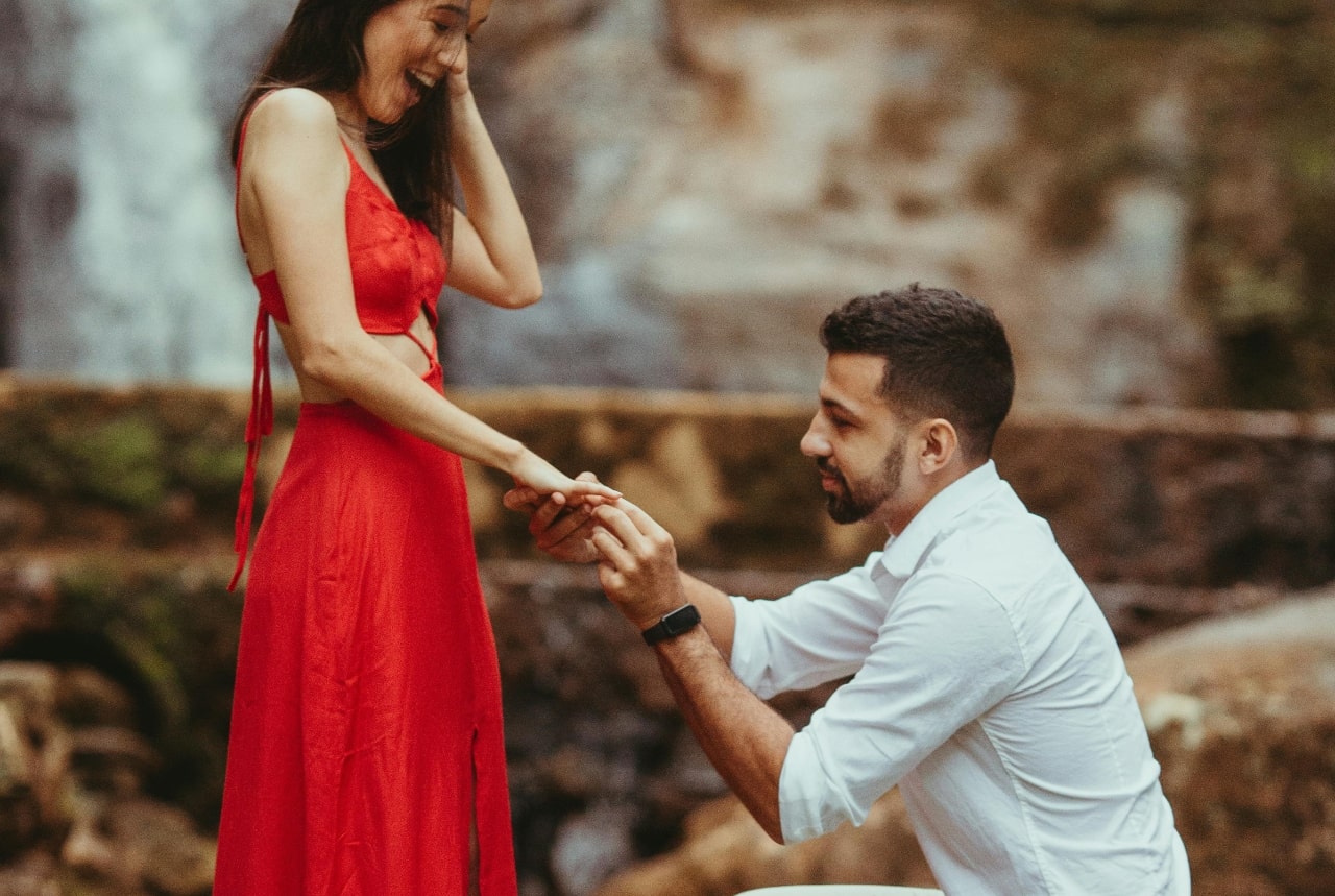A man on one knee, proposing to a woman in a bright red dress, in front of a beautiful nature-filled background.