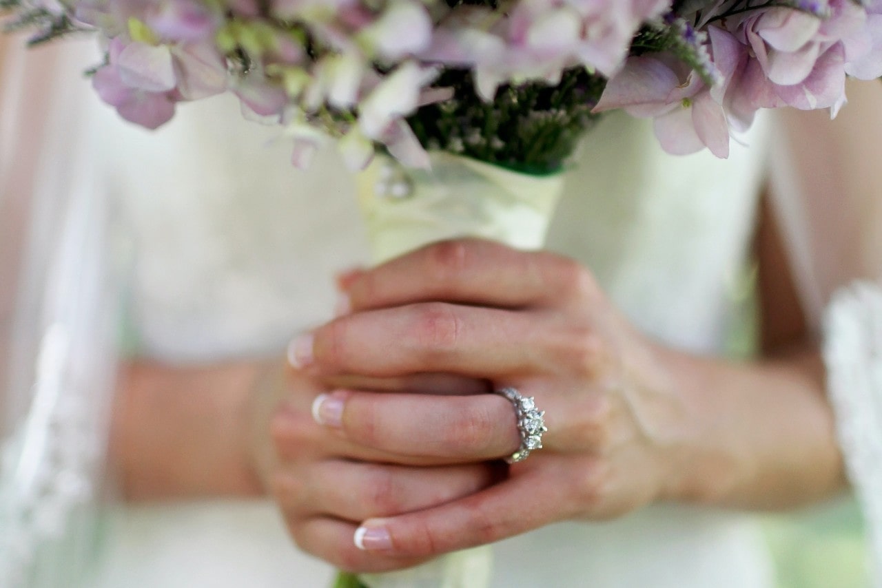 A close up of a bride's hand adorned with white gold three stone diamond engagement ring, while she holds a bouquet of flowers.