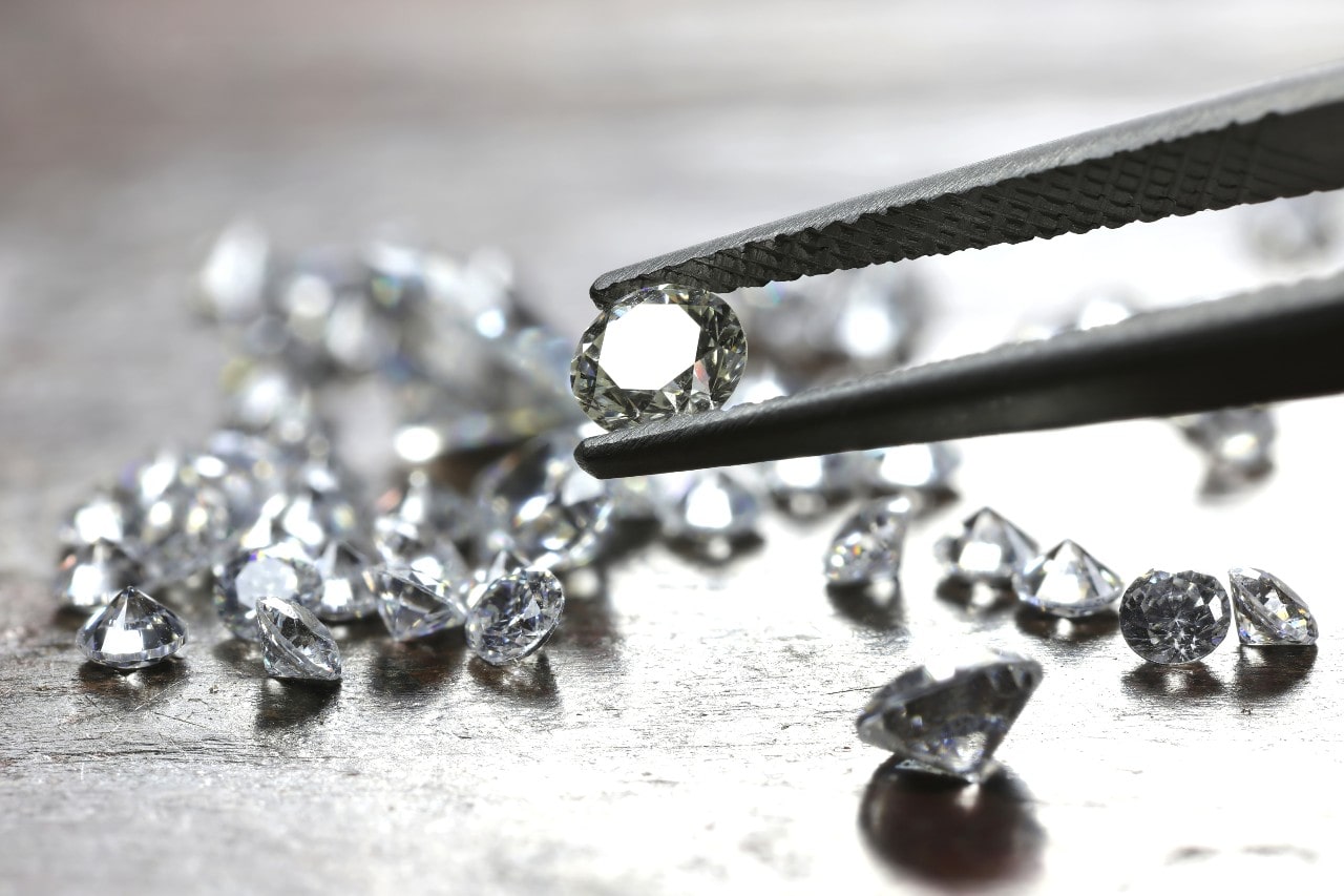 Tweezers hold a shiny diamond above a reflective surface, surrounded by scattered loose diamonds. Tweezers hold a shiny diamond above a reflective surface, surrounded by scattered loose diamonds.