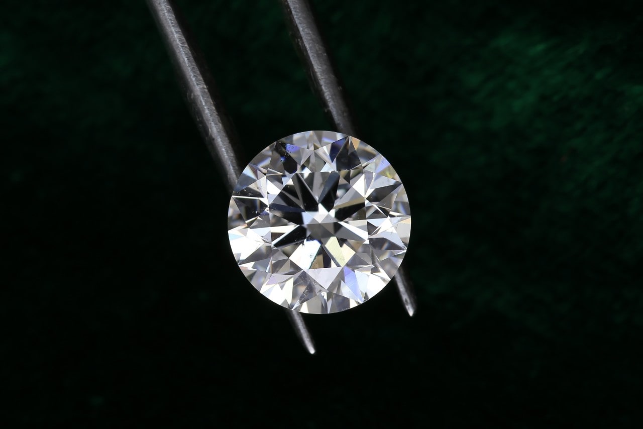 A sparkling, round-cut diamond is held by metal tweezers against a dark green, textured background, highlighting its clarity and brilliance. A sparkling, round-cut diamond is held by metal tweezers against a dark green, textured background, highlighting its clarity and brilliance.