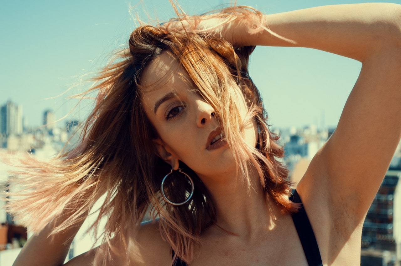 A woman with ginger hair and hoop earrings poses against a city landscape. A woman with ginger hair and hoop earrings poses against a city landscape.