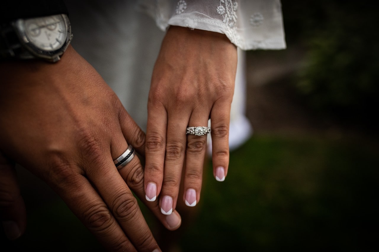 A close-up of a groom’s and bride’s hands, adorned with a silver wedding band and a side stone diamond engagement ring.