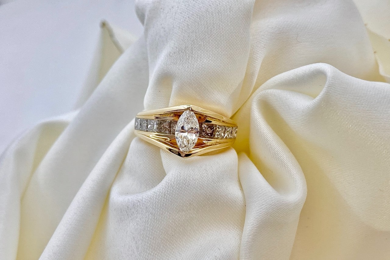 A yellow gold engagement ring with marquise cut diamond at the center and princess cut gemstones along the band on a white fabric.