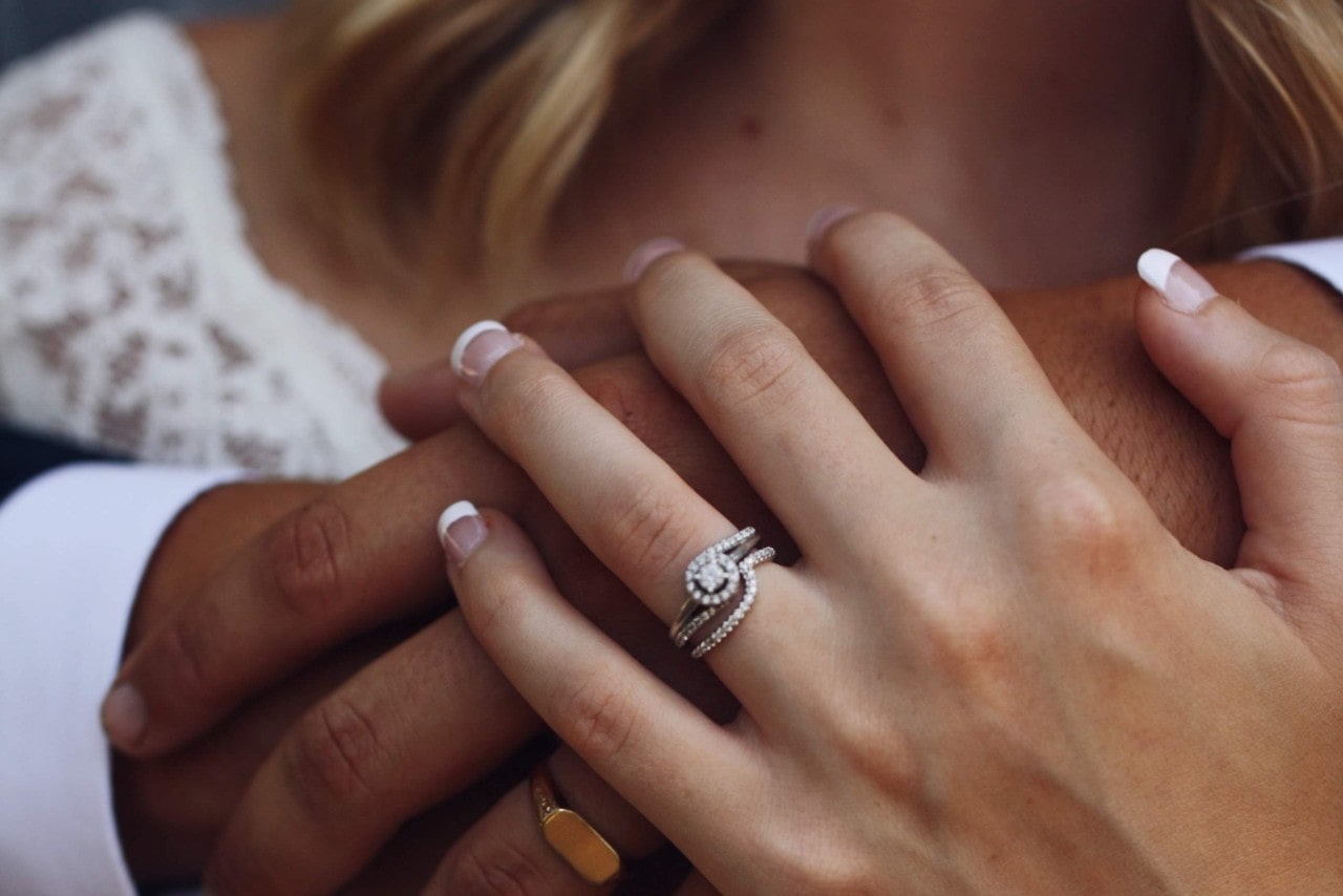 A close-up of a couple holding hands, showcasing woman’s diamond halo engagement ring.