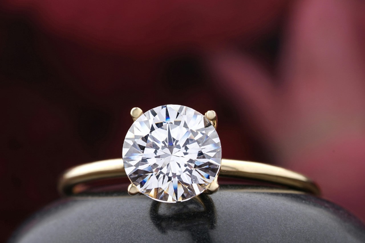 A yellow gold thin solitaire engagement ring with round cut diamond against a blurry burgundy background. A yellow gold thin solitaire engagement ring with round cut diamond against a blurry burgundy background.