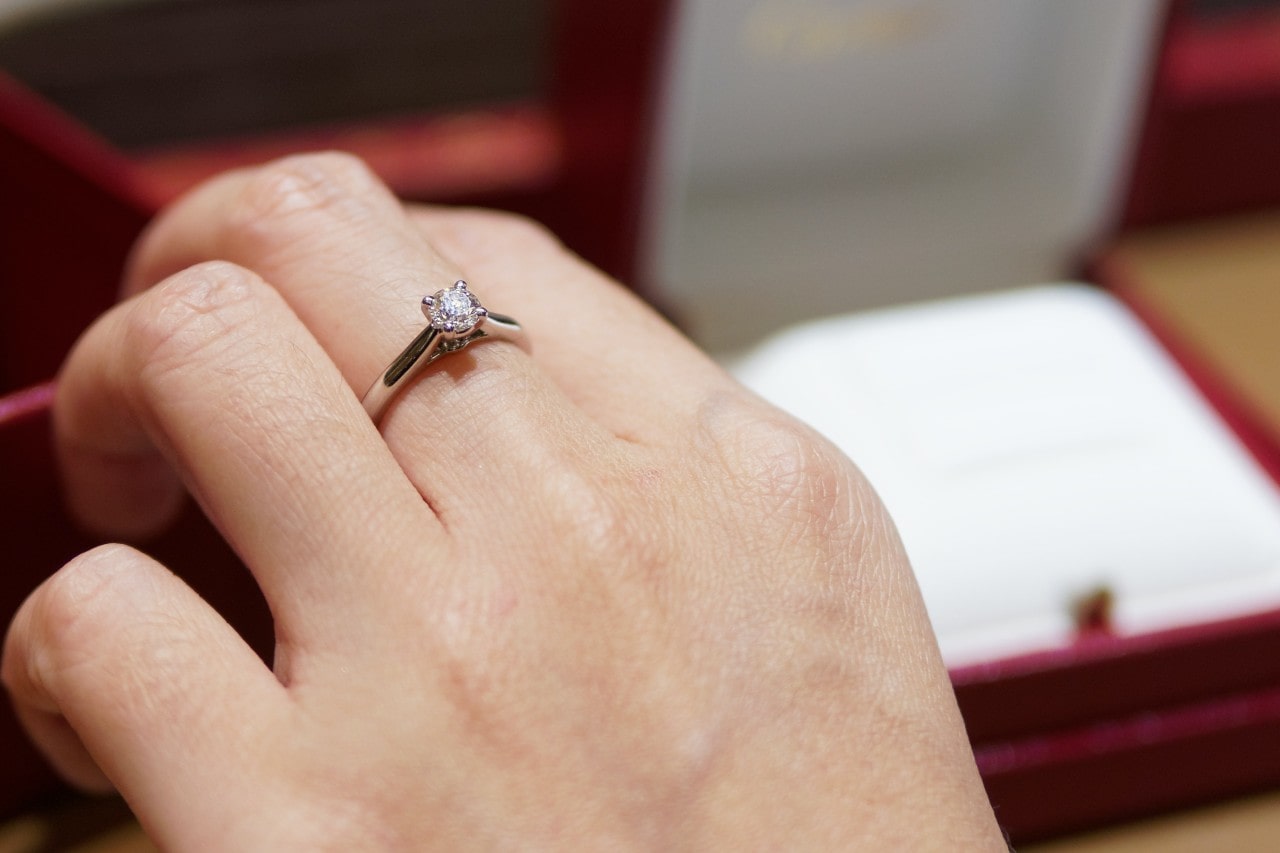 A close-up of a hand wearing a silver engagement ring with a single round diamond set in a prong setting. A close-up of a hand wearing a silver engagement ring with a single round diamond set in a prong setting.